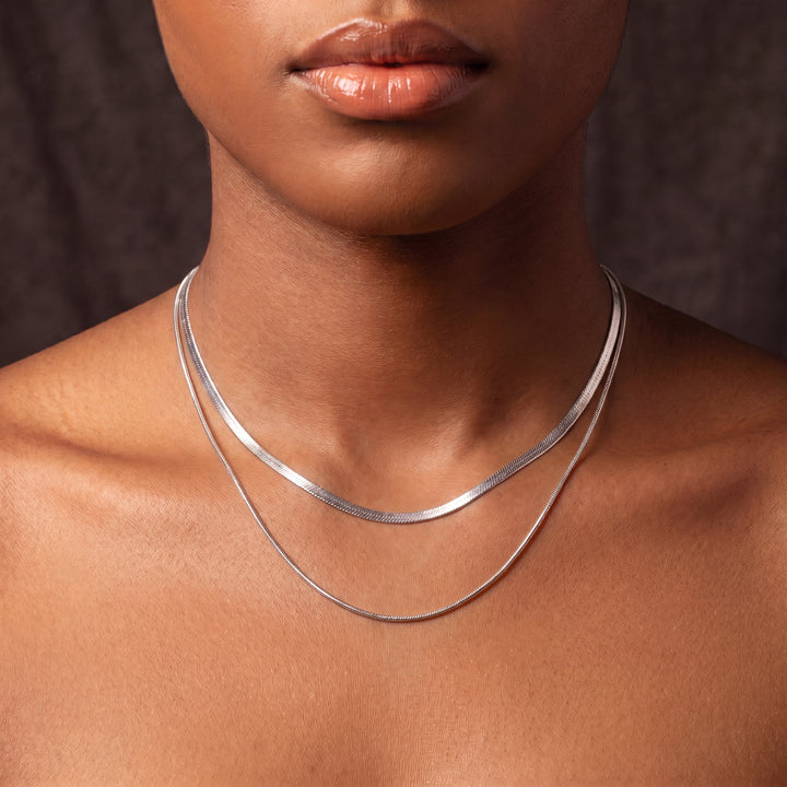 Layered silver necklaces on a model's neck, one flat snake chain, one thin snake chain.