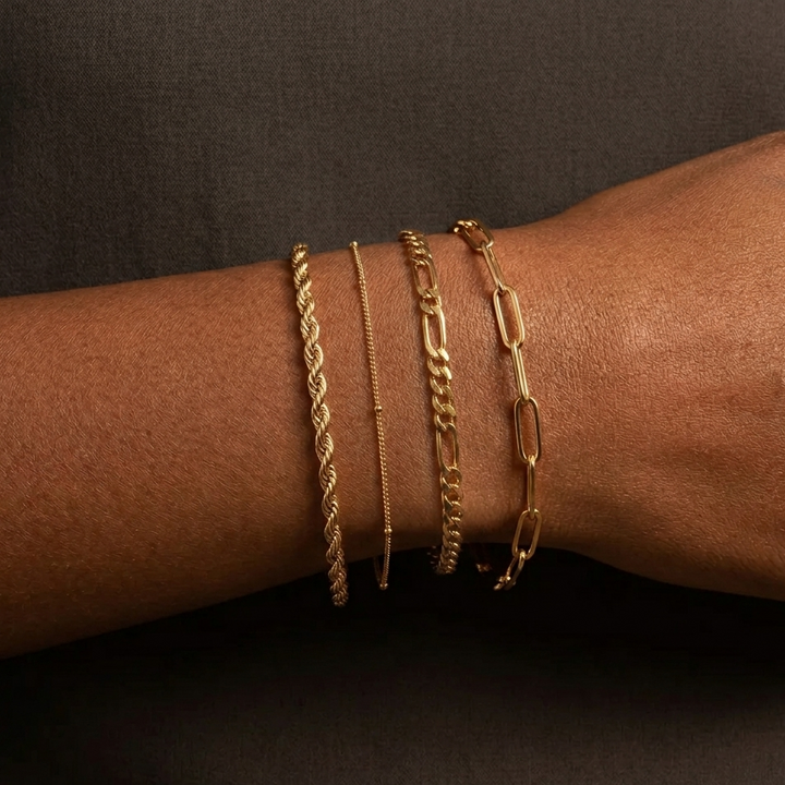 Four gold-toned bracelets: a rope, thin beaded, figaro, and paperclip chain, worn on a wrist.